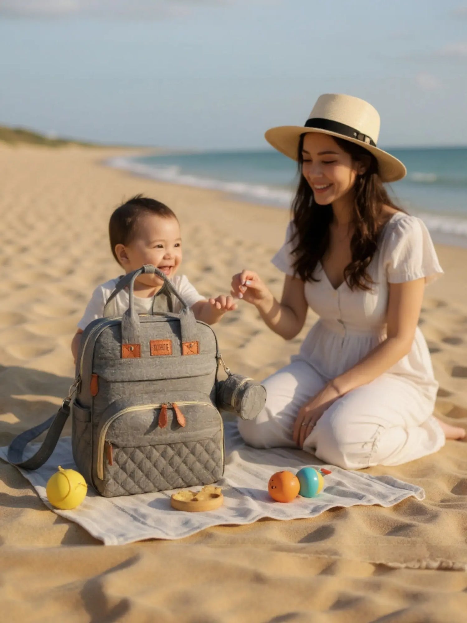 sac à dos à langer 3 en 1 gris posé sur la plage à côté d’une femme et de son bébé