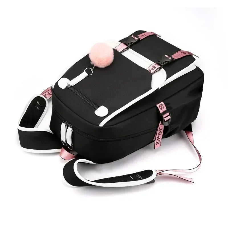 Sac à dos scolaire noir et rose avec port USB et pompon décoratif, posé à plat.