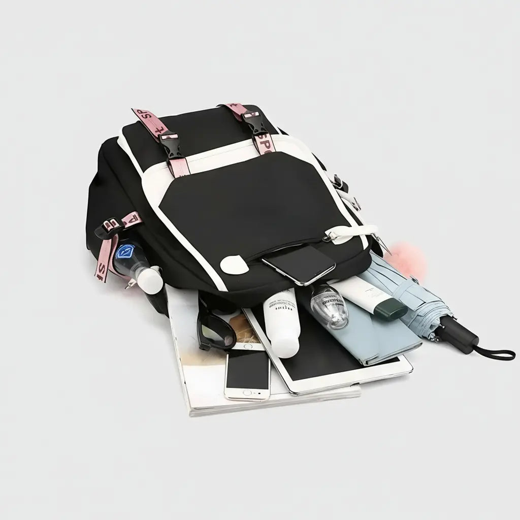 Sac à dos scolaire noir et rose avec port USB et pompon décoratif, ouvert et rempli de fournitures scolaires et électroniques.