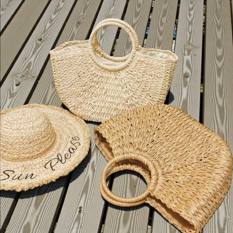 Sac en paille forme coquillage avec anses rondes rigides posé au sol à côté d’un chapeau