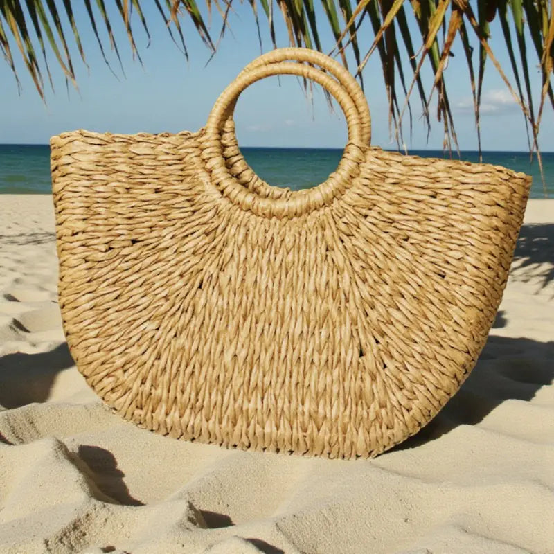 Sac en paille forme coquillage avec anses rondes rigides posé sur le sable au bord de la mer